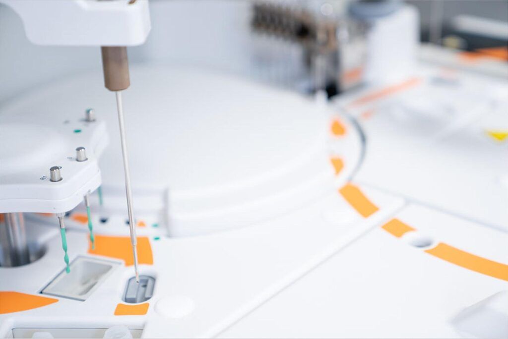 Close-up of a laboratory device with a robotic arm and needle poised over a sample holder, crucial for arthritis research. The equipment features a clean, white and orange design, indicative of modern lab technology.