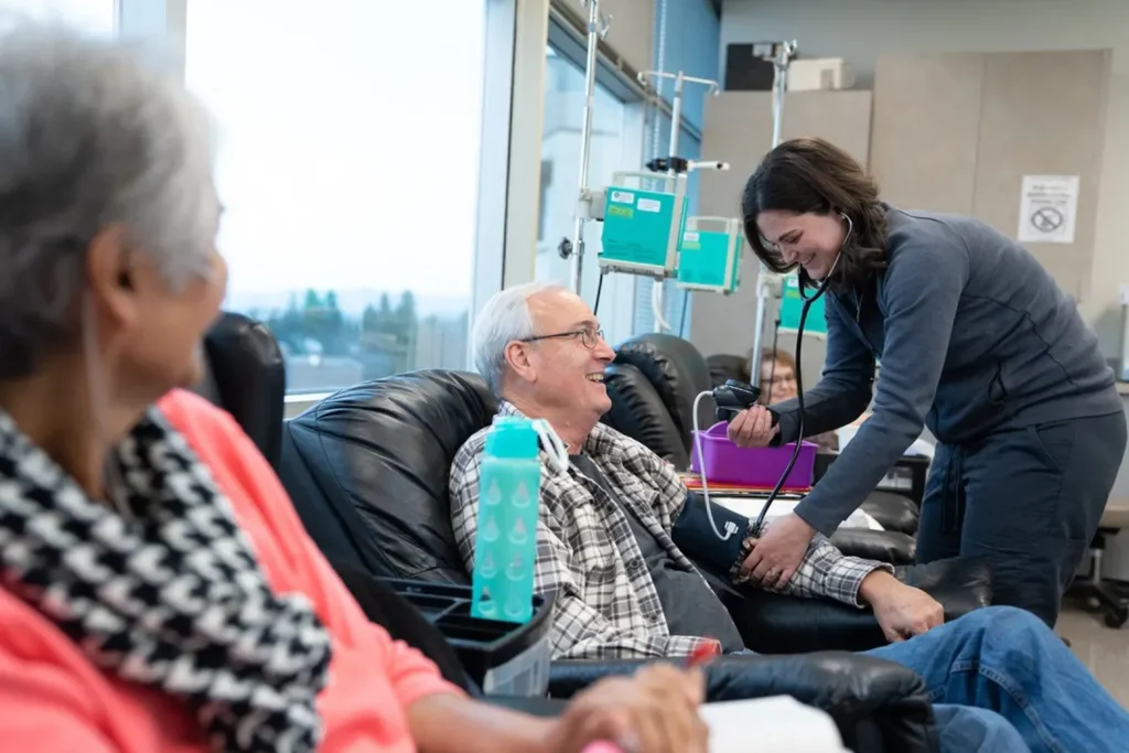 In a medical facility filled with equipment, a healthcare worker takes the blood pressure of an older man seated and smiling. Nearby, another person in a scarf observes attentively, reflecting the center's dedication to arthritis research.